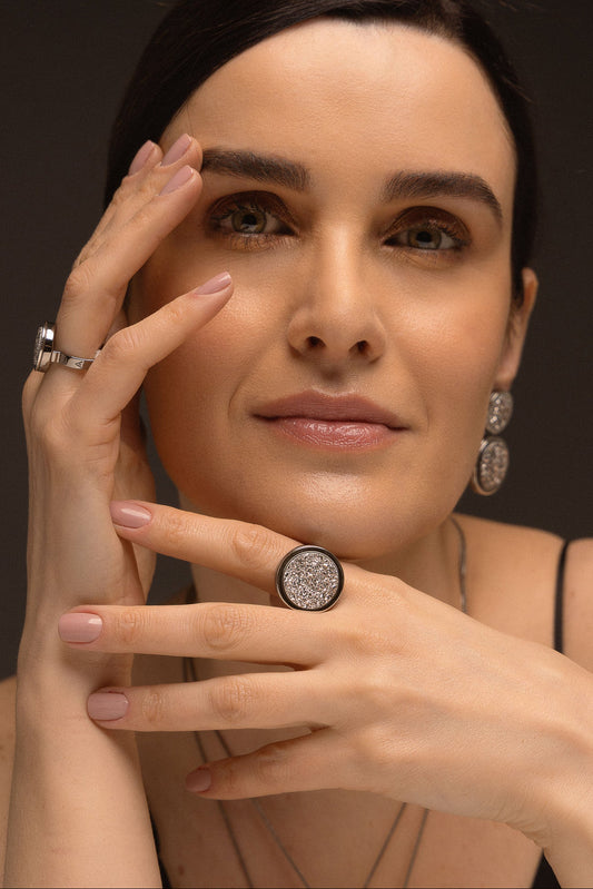 O Anel Único - Drusa Platina, da Coleção Singular, é uma peça de impacto que une o rústico ao sofisticado. A Drusa Platina é conhecida por sua capacidade de amplificar energias positivas, promover o equilíbrio e trazer brilho pessoal. Banhado em Ródio Negro, este anel apresenta um contraste moderno e elegante, destacando a beleza bruta da pedra em um design imponente que celebra a individualidade feminina.
