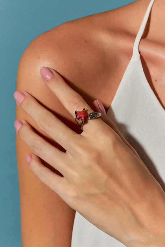 O Anel Serena com Feldspato Vermelho em Banho de Ródio Negro possui duas estrelas-do-mar que se encontram na mesma direção, simbolizando harmonia e união. A cor intensa do Feldspato Vermelho, em contraste com o brilho escuro e metálico do Ródio Negro, cria um visual sofisticado e marcante.