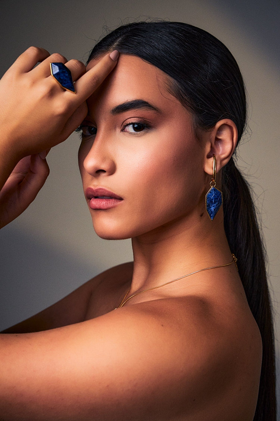 Modelo com a mão na testa usando o Brinco Impulso com Sodalita Azul, brinco comprido e anguloso banhado a ouro 18k